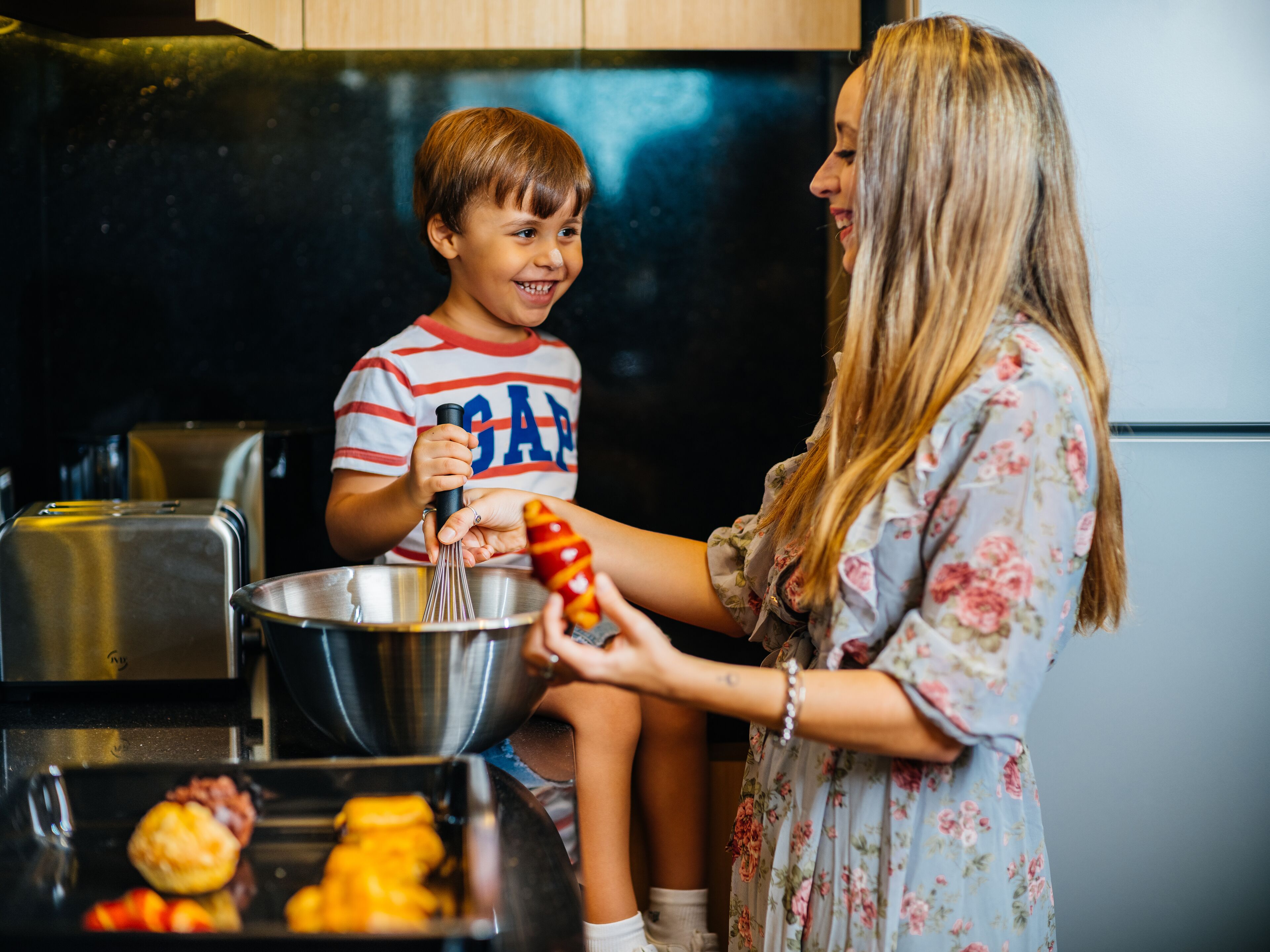 Una cucina con elettrodomestici in acciaio inossidabile, un bambino che tiene una frusta e una donna con un abito floreale.