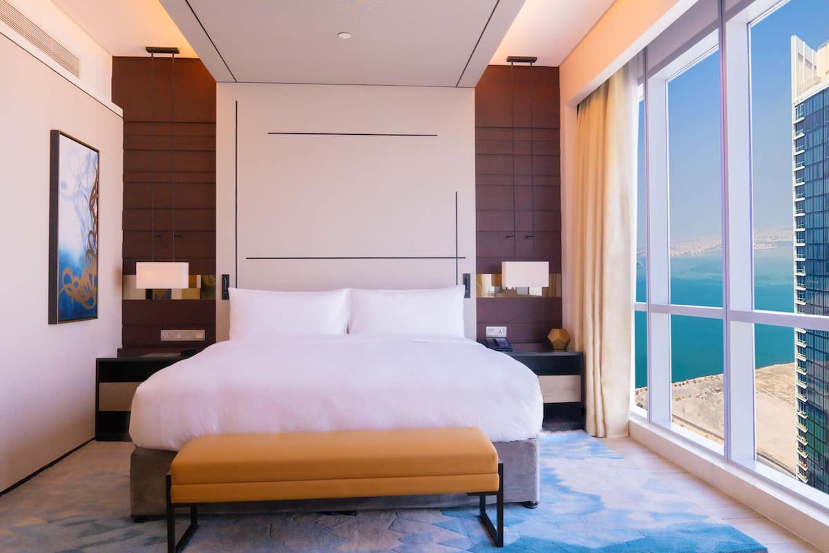 A modern hotel room with a large bed, a bench, a nightstand with a lamp, and a view of the city through large windows.