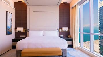 A modern hotel room with a large bed, a bench, a nightstand with a lamp, and a view of the city through large windows.