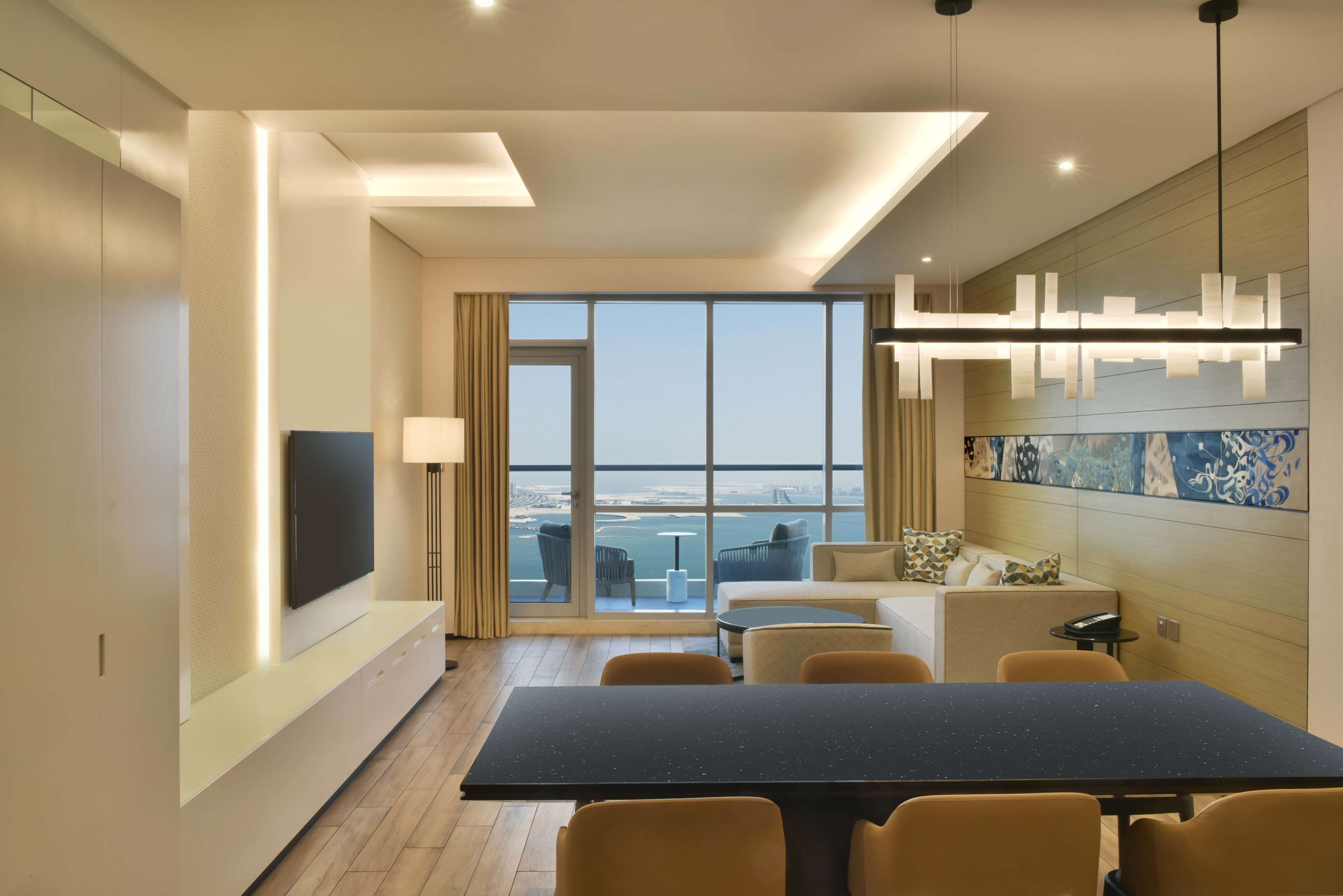 A modern hotel room with a large window offering a city view, a flat-screen TV mounted on the wall, a dining area with a table and chairs, and a decorative light fixture.