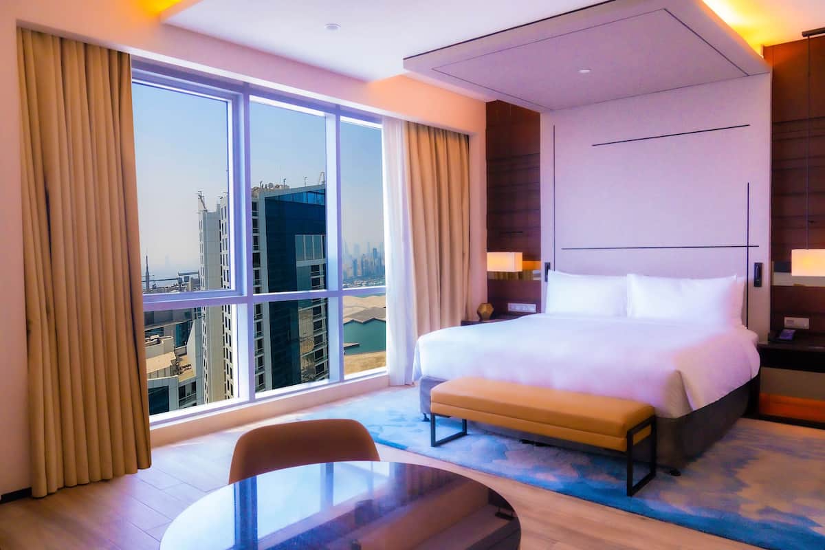 A hotel room with a large bed, a bench, a table, and a view of the cityscape through large windows.