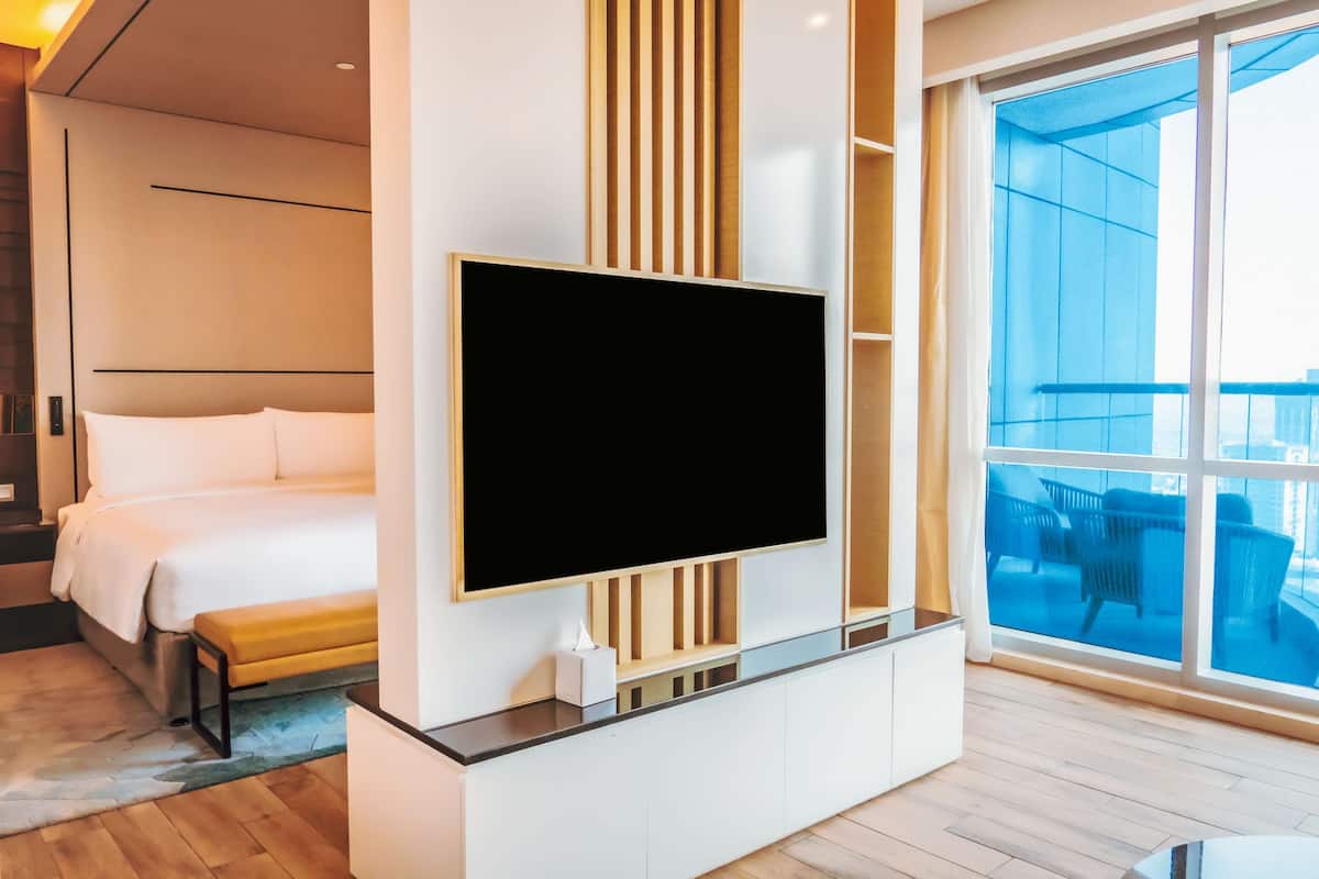A modern hotel room with a large flat-screen TV mounted on a wall, a bed with white linens, a wooden bench, and a balcony with outdoor furniture.