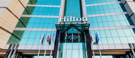 The exterior of a Hilton hotel with a modern glass facade, multiple flags, and the hotel's name prominently displayed.