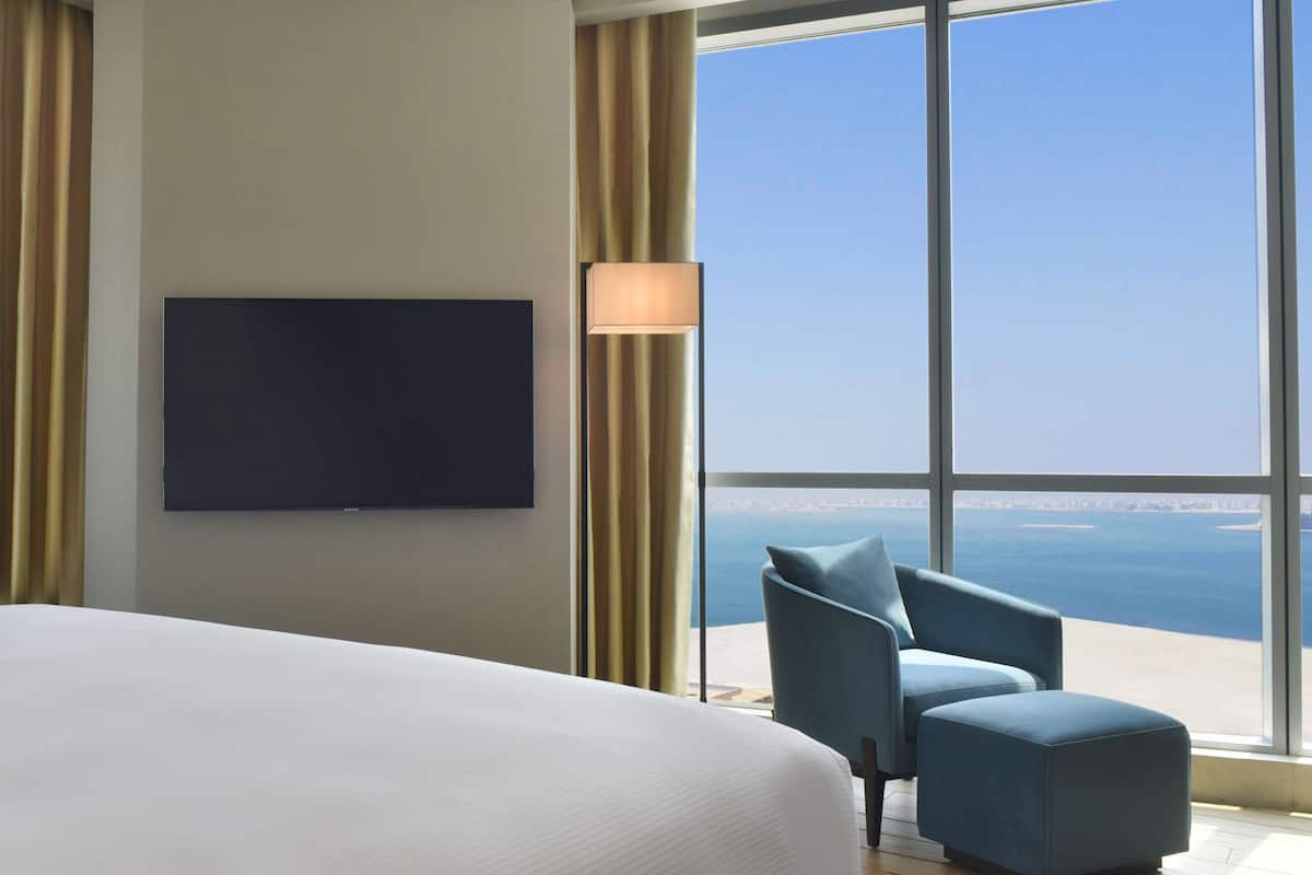 A hotel room with a large bed, a flat-screen TV mounted on the wall, a blue armchair, and a view of the sea through large windows.