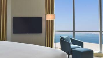 A hotel room with a large bed, a flat-screen TV mounted on the wall, a blue armchair, and a view of the sea through large windows.