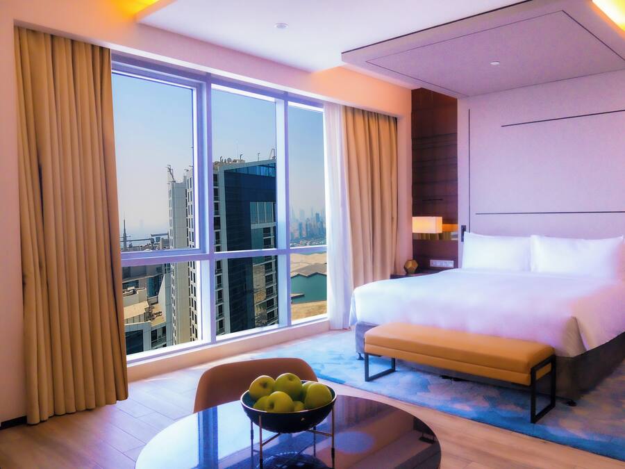 Habitación de hotel con una cama grande, una mesita de noche, una silla, un cuenco con fruta y vistas a la ciudad a través de amplias ventanas.
