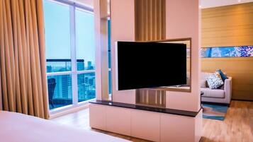 Een moderne hotelkamer met een grote televisie bevestigd aan een witte kast, een groot raam met gordijnen, een bank met gebogen kussens en uitzicht op de skyline.
