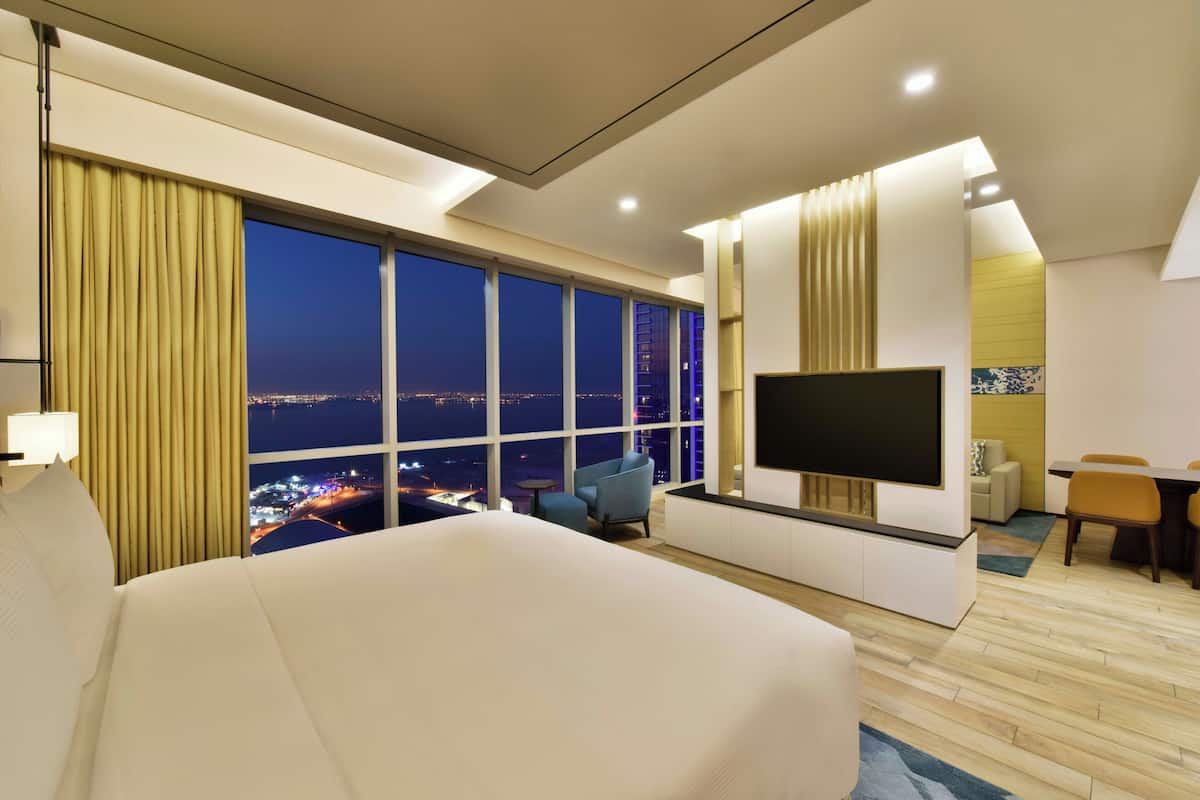 A hotel room with a large bed, a TV, a desk, and a view of the city at night.