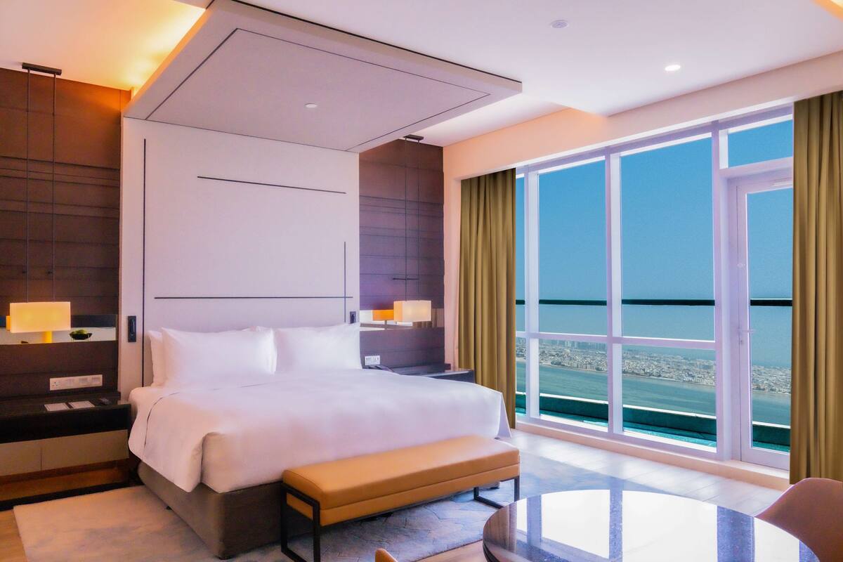A modern hotel room with a large bed, bedside tables, a bench, and a view of the city through large windows.