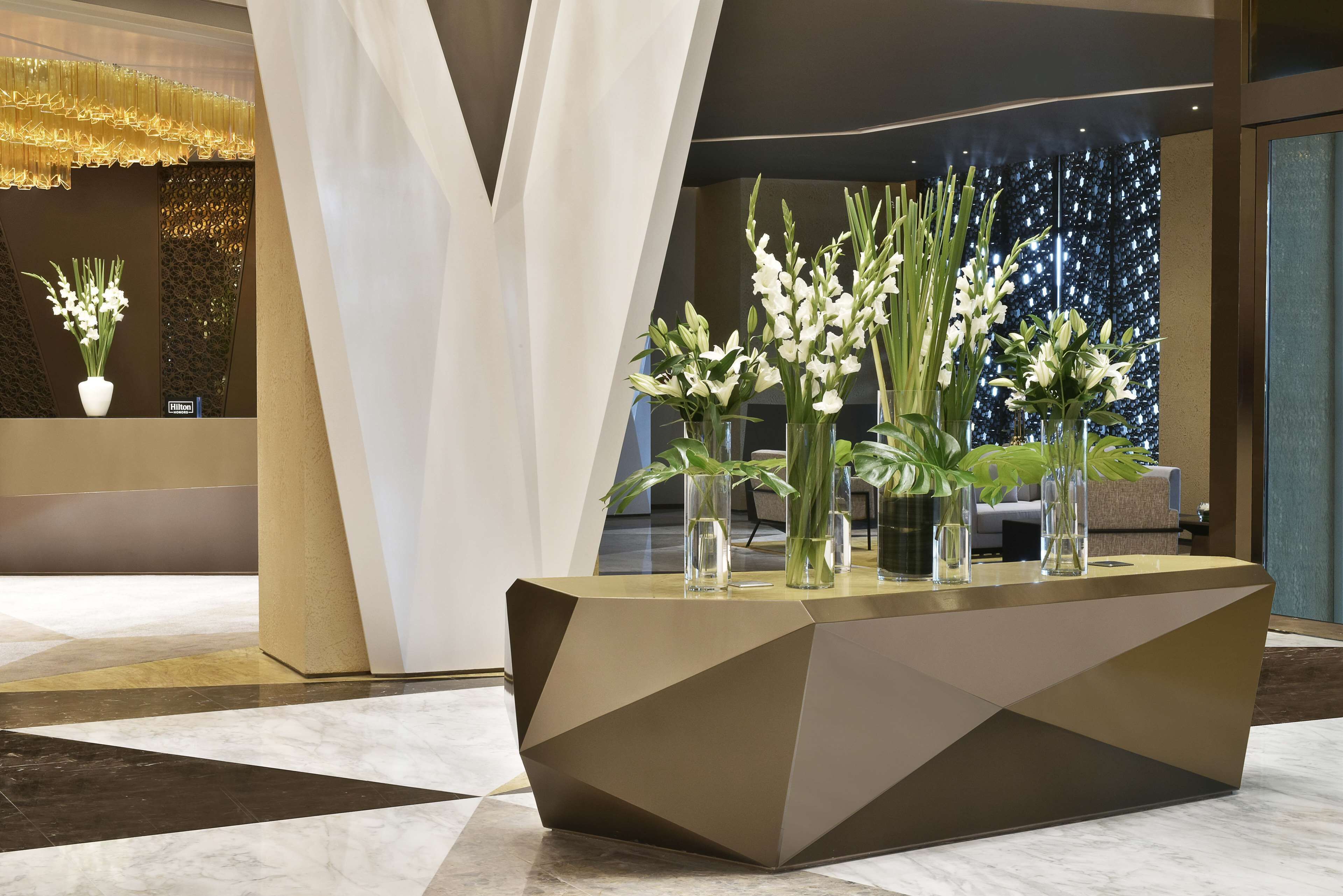 A reception desk with a geometric design, surrounded by vases of white flowers, in a modern lobby with a marble floor and a decorative wall.