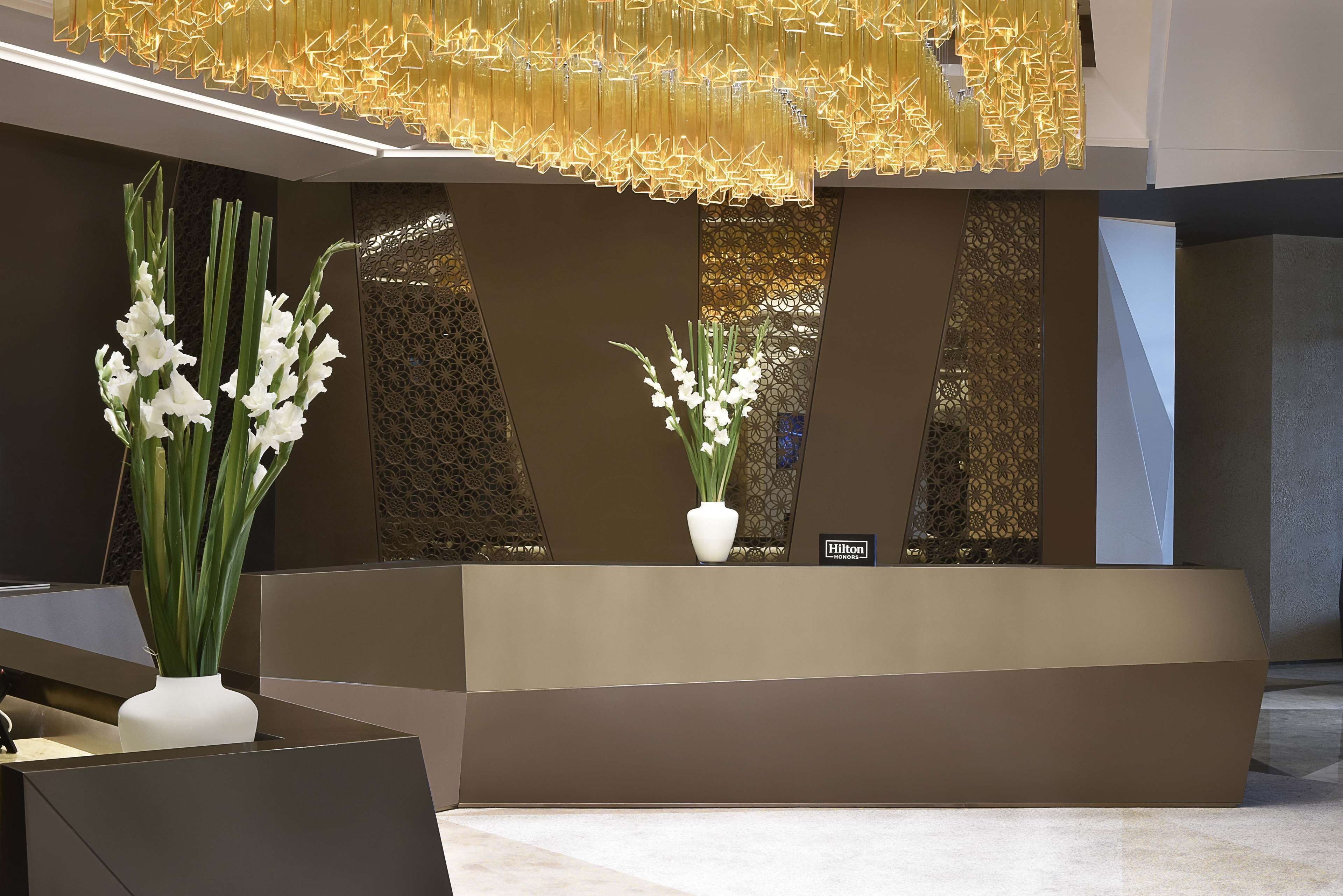 A modern reception desk with two white vases of flowers, a Hilton sign, and a decorative golden light fixture above.