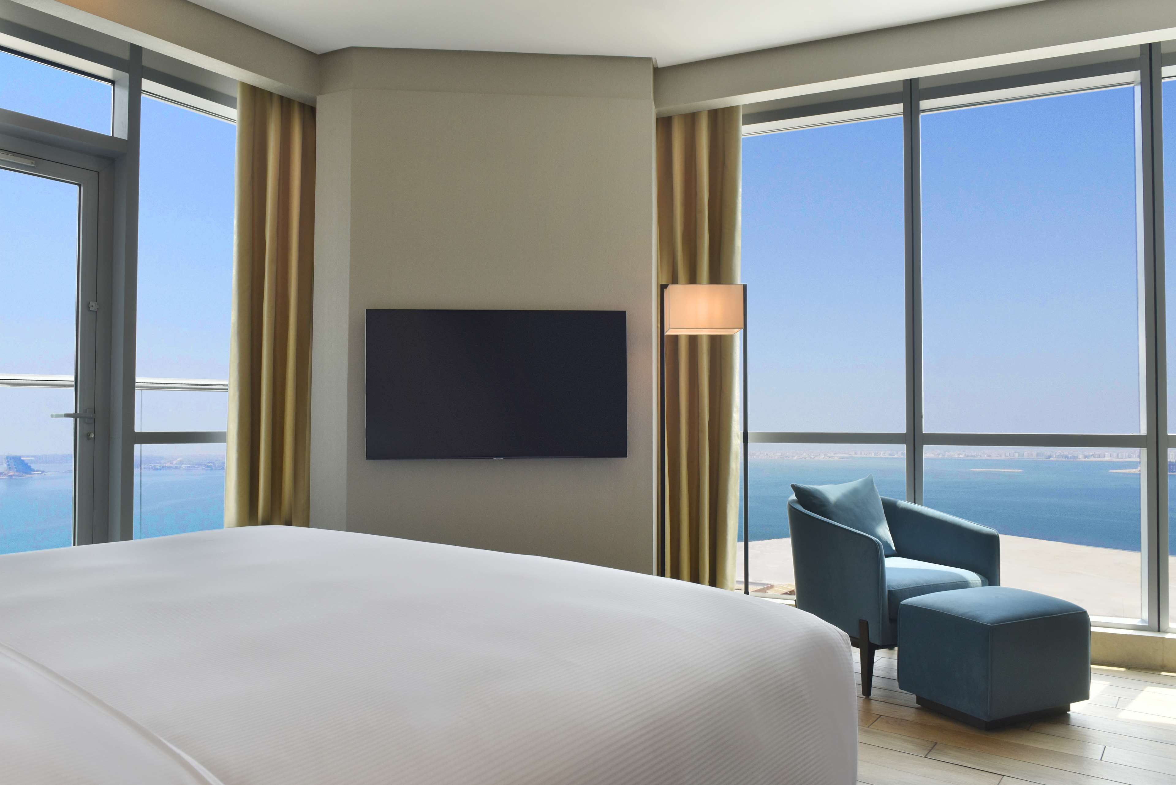 Habitación de hotel con una cama grande, un televisor de pantalla plana montado en la pared, una silla y vistas al mar a través de amplias ventanas.