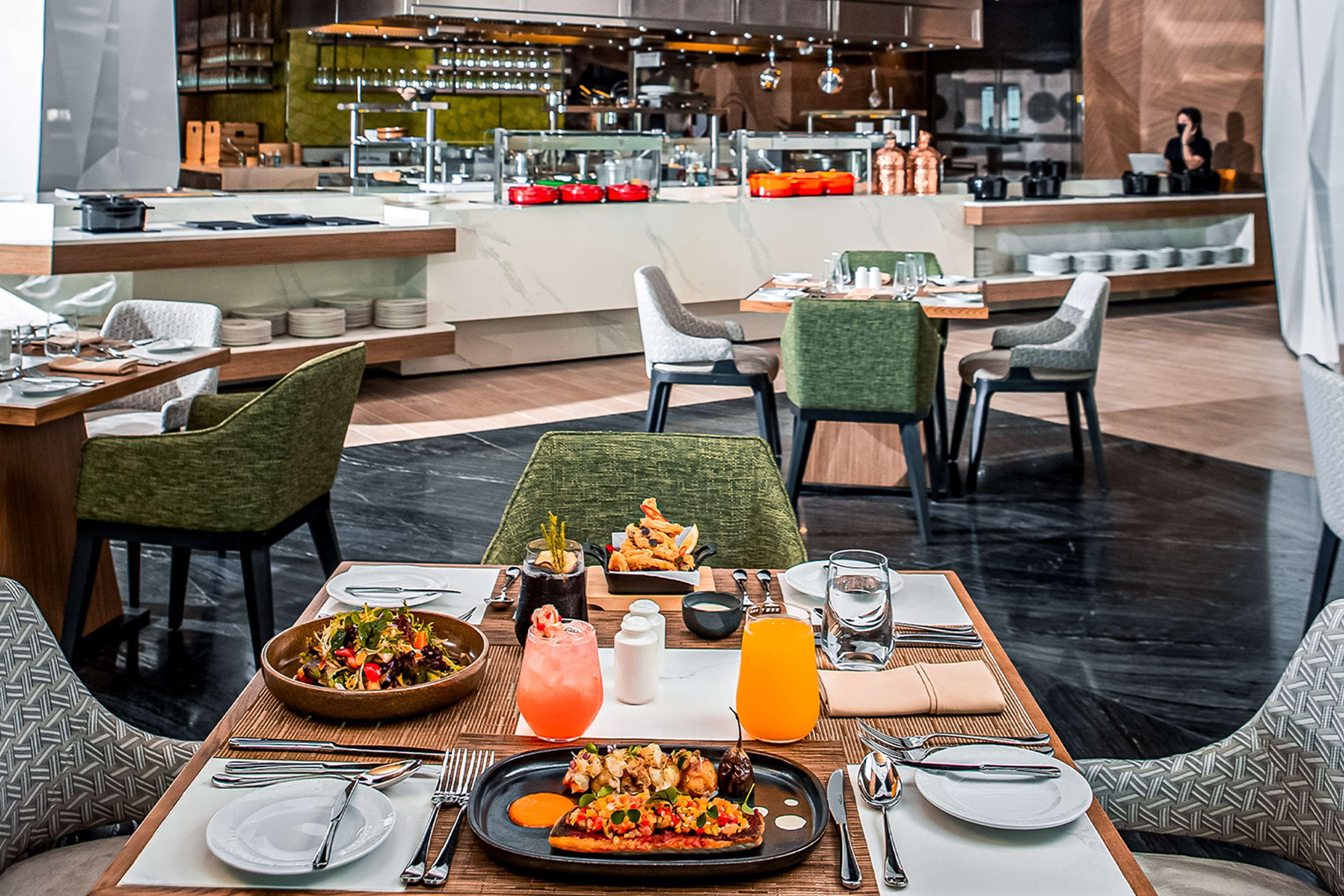 A restaurant with a modern interior, featuring a central dining table with a variety of dishes, including a large platter of food, a bowl of salad, and a glass of orange juice. There are multiple chairs around the table, and the background shows a bar area with a bartender.