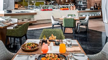 A restaurant with a modern interior, featuring a central dining table with a variety of dishes, including a large platter of food, a bowl of salad, and a glass of orange juice. There are multiple chairs around the table, and the background shows a bar area with a bartender.