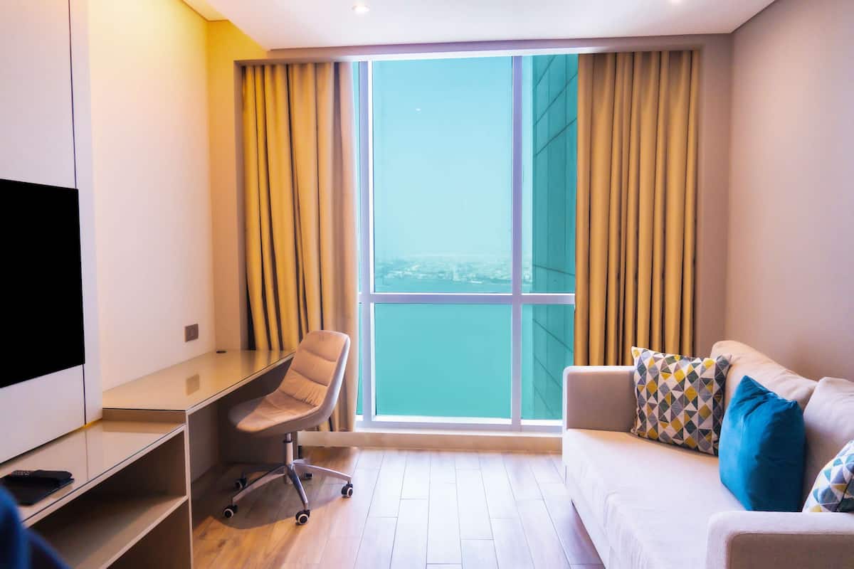 A modern hotel room with a large window, a desk, a chair, a sofa, and a TV.