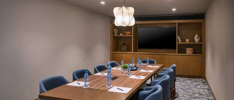 Un salón de conferencias con una larga mesa de madera, sillas azules, botellas de agua y una pantalla grande en la pared.