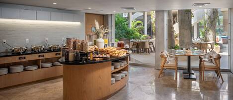 A modern cafe with a central counter displaying pastries and beverages, surrounded by seating areas with natural light and views of greenery.