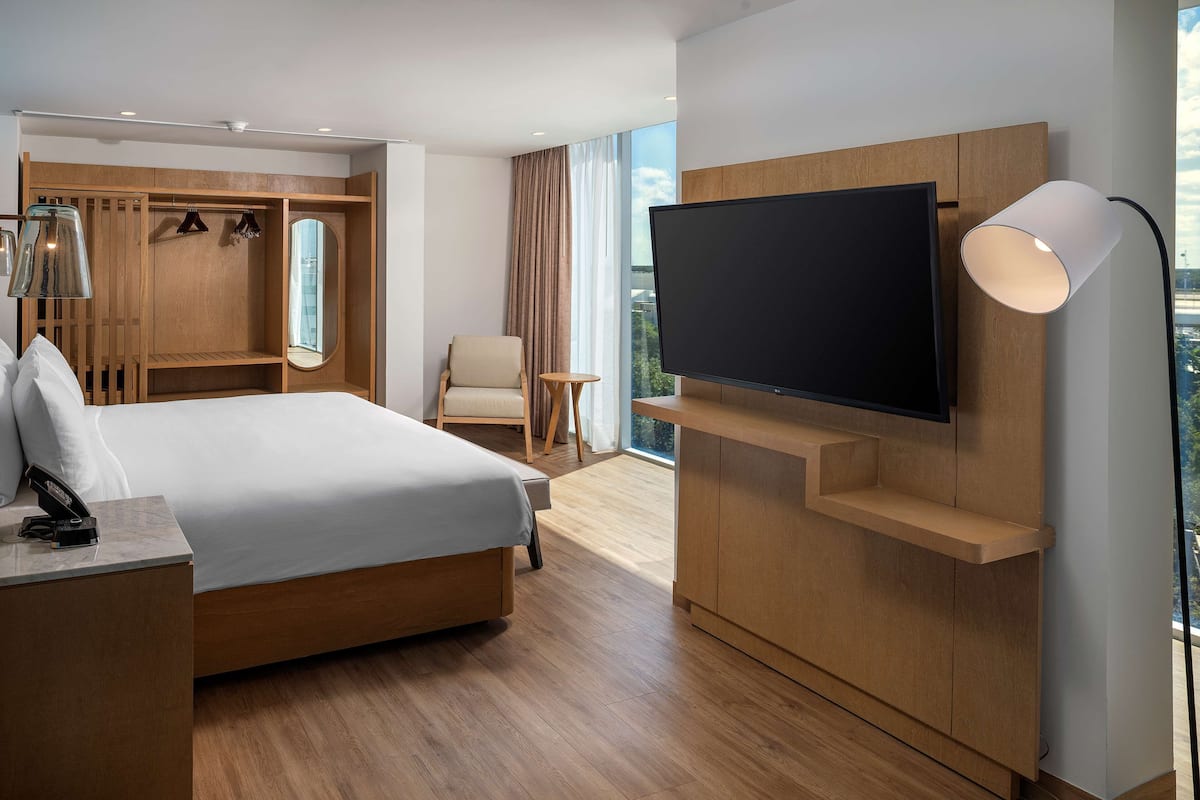 A modern hotel room with a large bed, a TV mounted on the wall, a desk with a lamp, a wardrobe, and a view of the city.