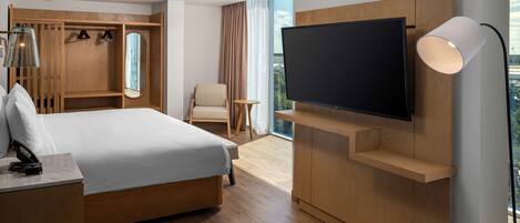 Quarto de hotel moderno com uma cama grande, televisão na parede, escrivaninha com luminária, guarda-roupa e vista para a cidade.