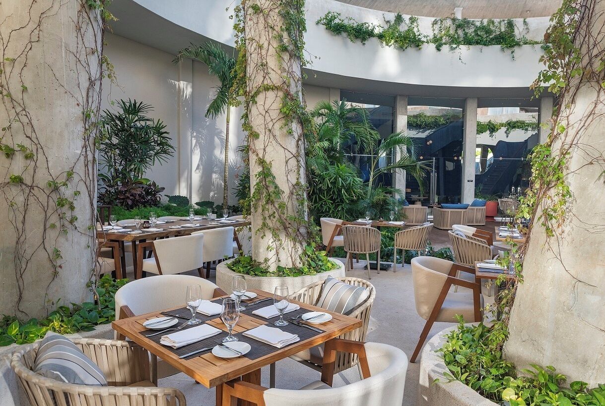 An outdoor dining area with tables and chairs, surrounded by greenery and columns.