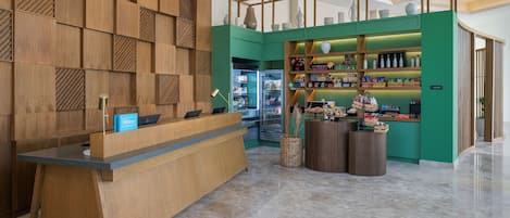 A hotel reception area with a wooden reception desk, a green counter with shelves, and a refrigerator in the background.