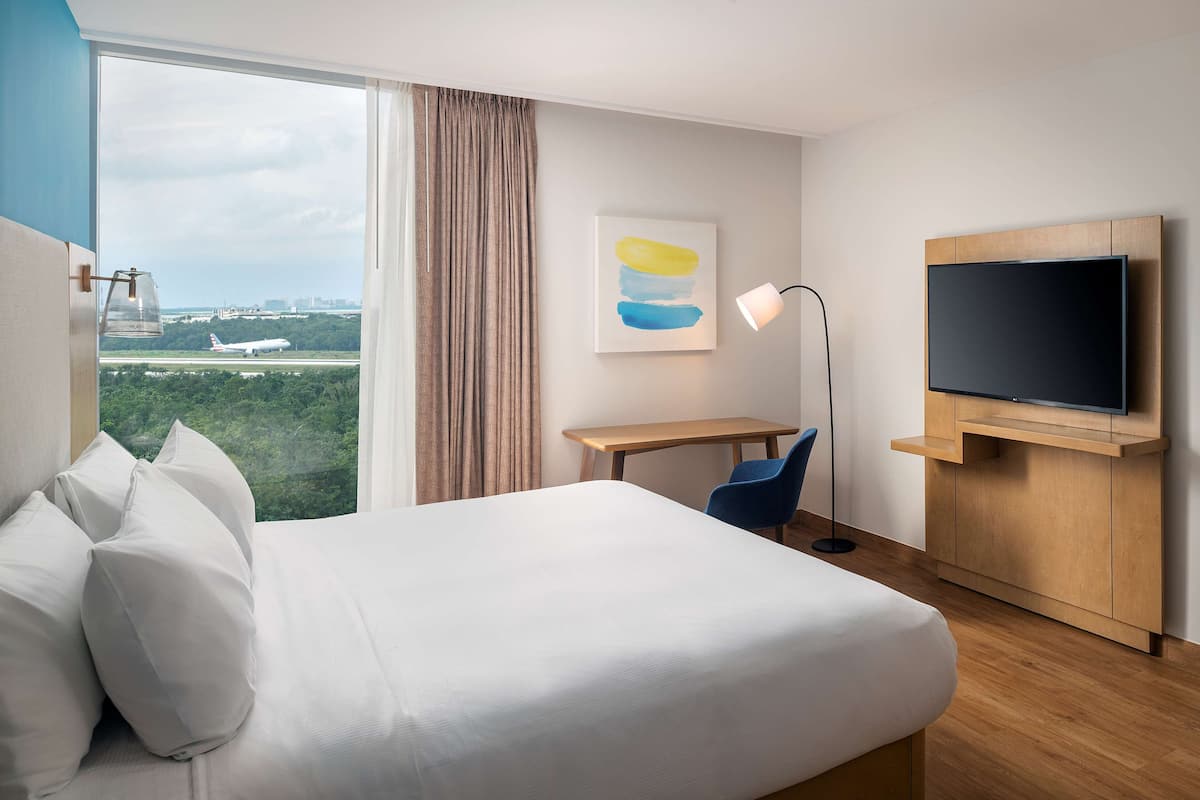 A hotel room with a bed, a desk, a chair, a TV, a lamp, and a window with a view of an airplane and greenery.