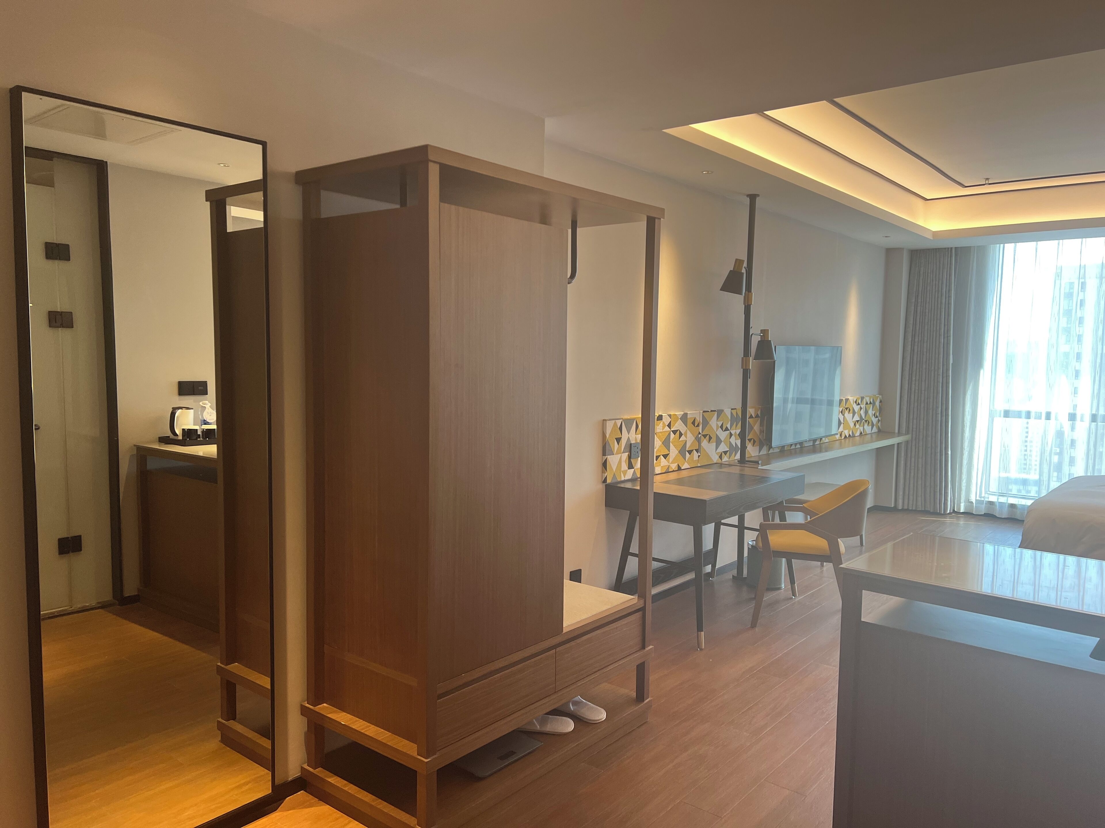 Een moderne hotelkamer met een houten garderobe, een bureau met stoel, een grote spiegel en uitzicht op de stad vanuit het raam.
