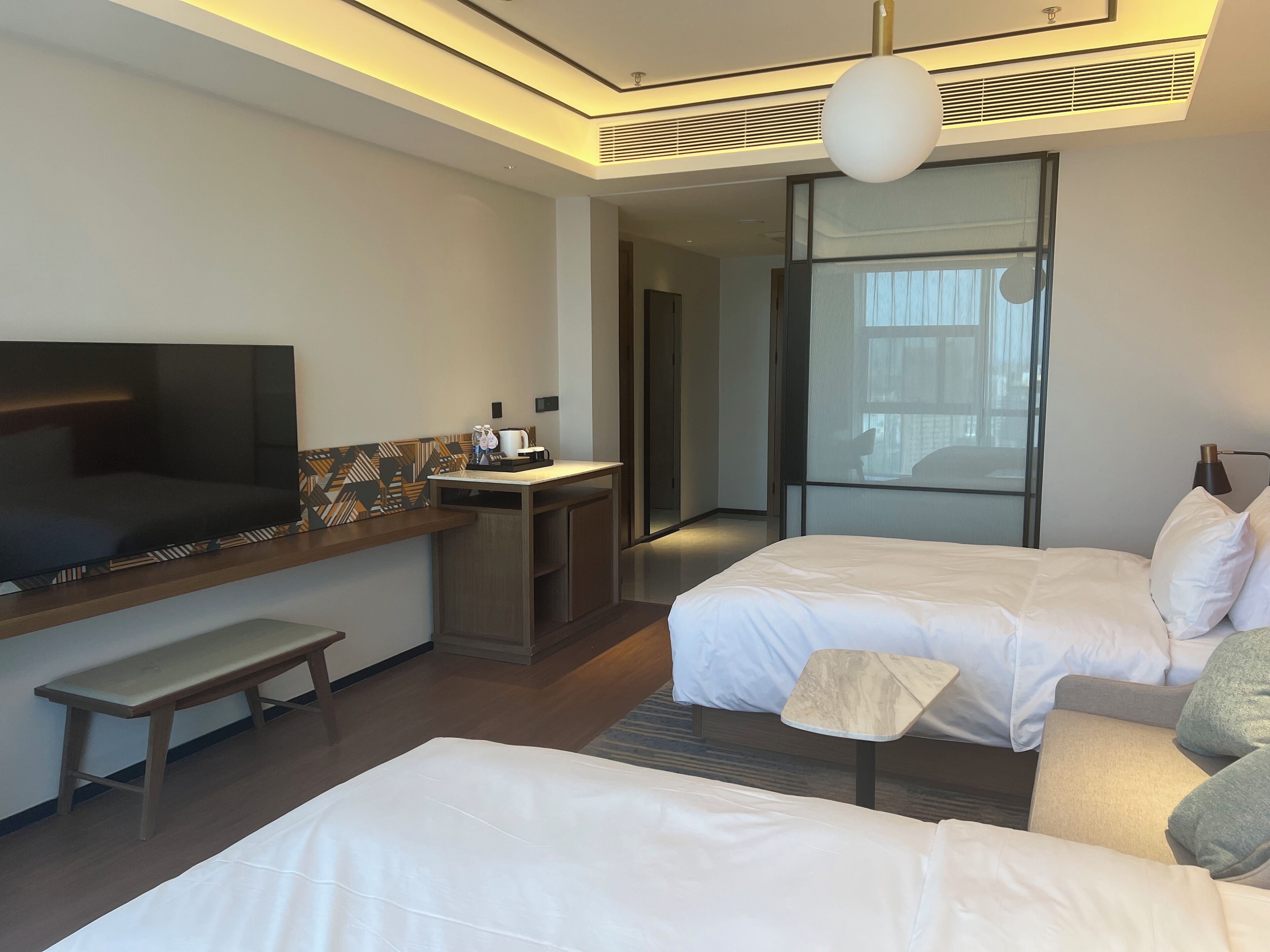 A hotel room with two beds, a TV, a small desk with a coffee maker, and a bench.
