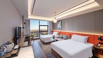 A modern hotel room with a large bed, a desk with a chair, a small table with a vase, and a view of the city through large windows.
