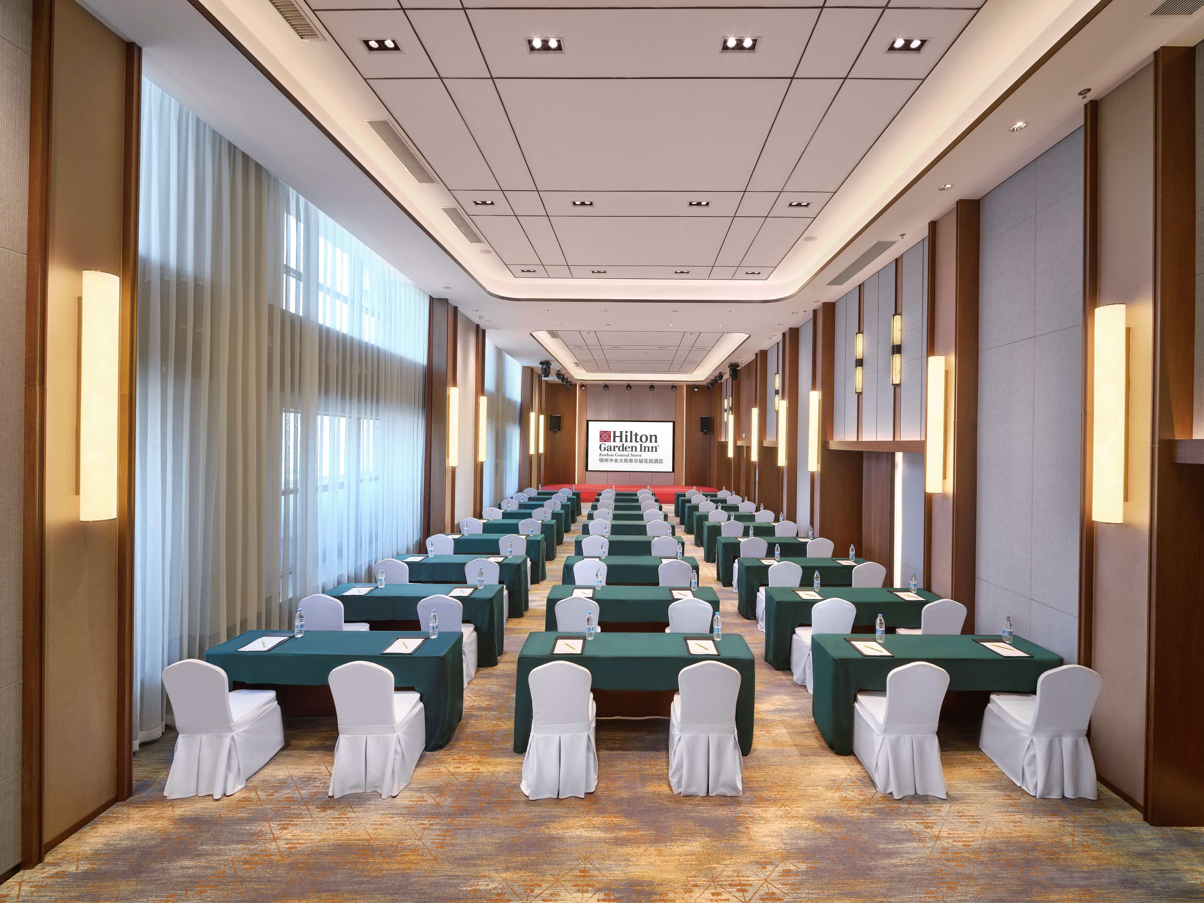 Ett konferensrum med rader av bord och stolar, en stor skärm som visar Hilton Garden Inns logotyp och ett golv täckt av matta.