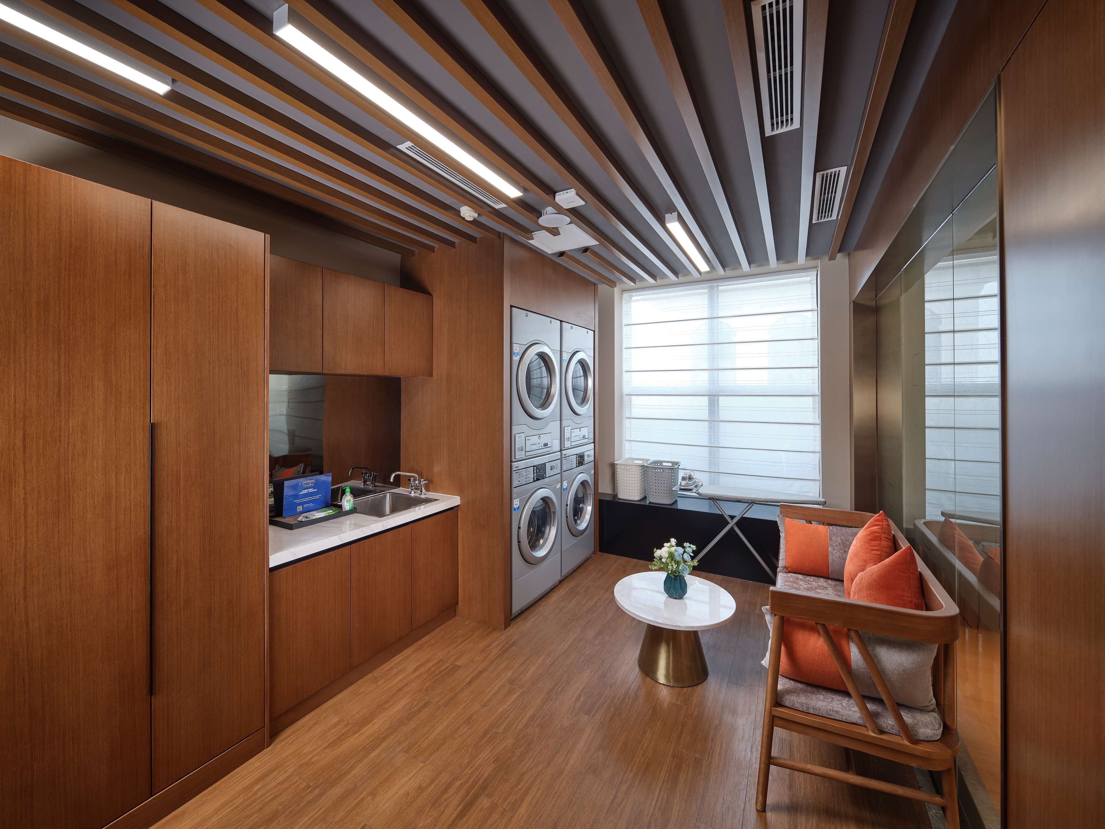 A compact kitchen with wooden cabinets, a built-in washing machine, a small dining table, and a window with blinds.