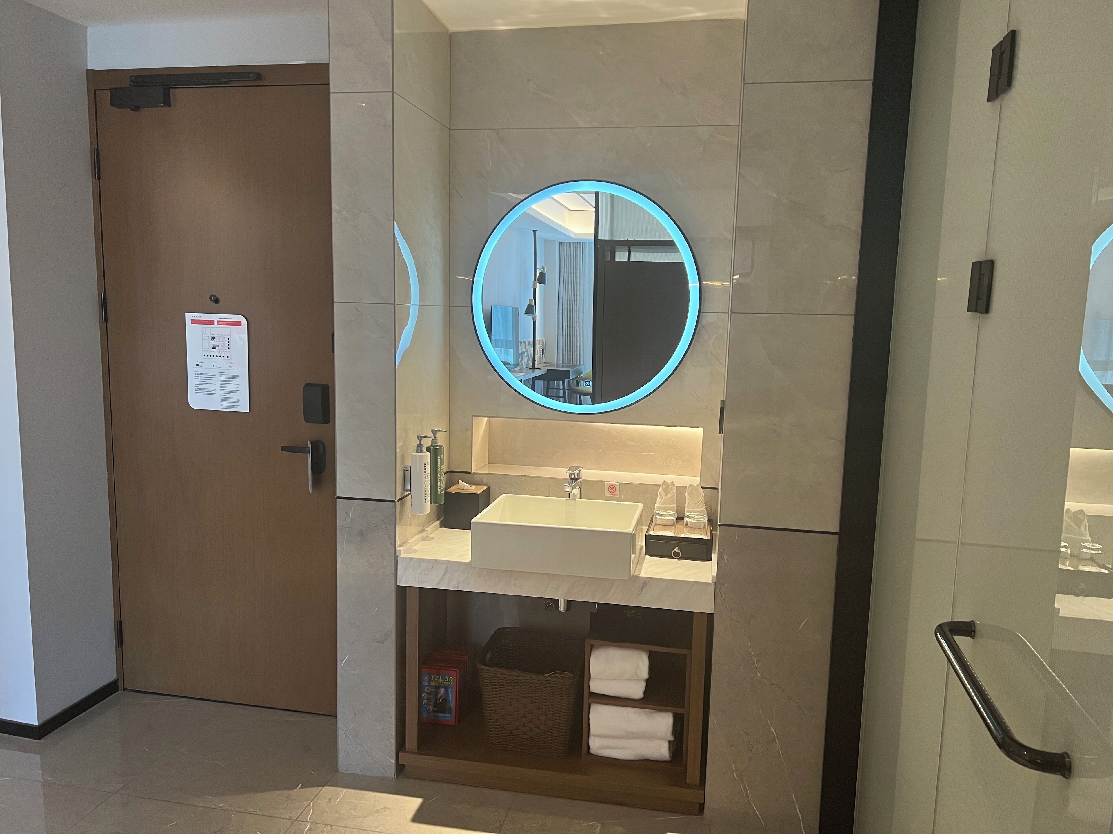 Un bagno moderno con una porta in legno, uno specchio rotondo con cornice blu, un lavabo e uno scaffale con asciugamani.