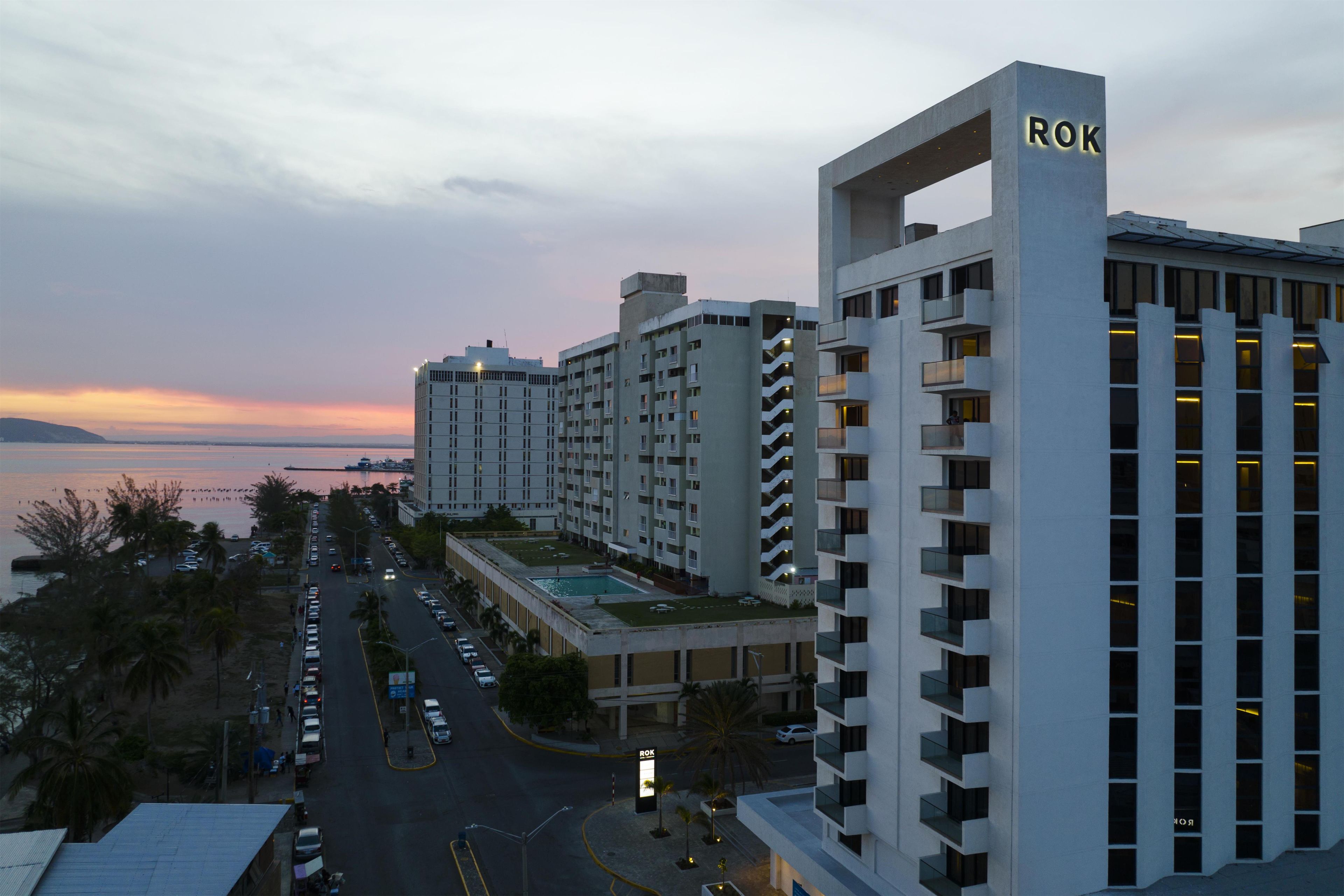 A high-rise hotel with a rooftop pool, multiple balconies, and a view of the sea at dusk.