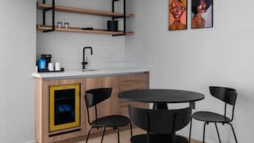 Uma cozinha moderna com prateleiras de madeira, uma torneira preta, uma máquina de café e um armário amarelo com porta preta. Há duas cadeiras pretas ao redor de uma mesa redonda.