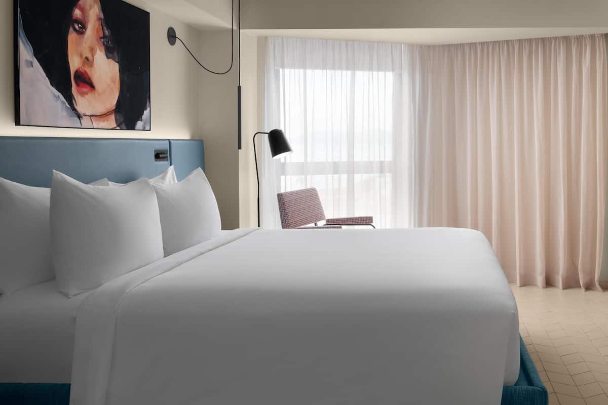 A hotel room with a large bed, a chair, a desk, a lamp, and a painting on the wall.