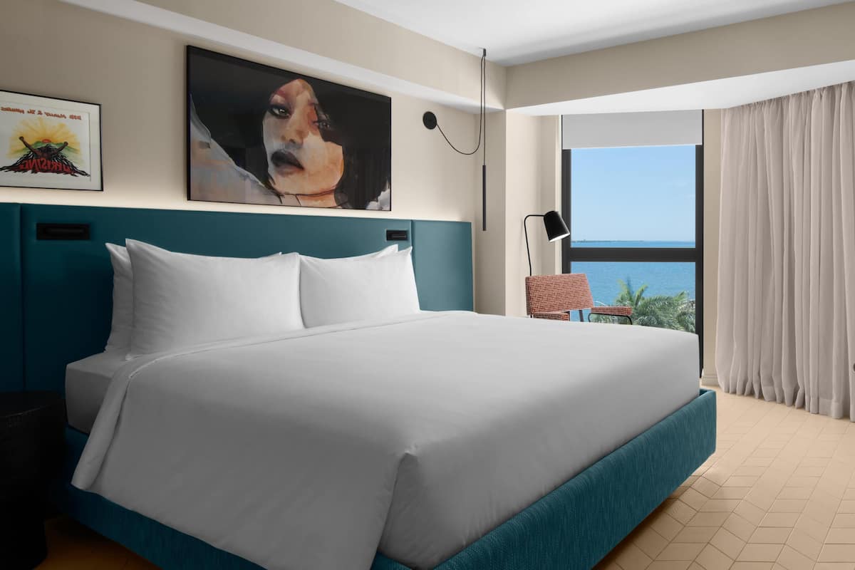 A hotel room with a large bed, a view of the ocean, and artwork on the wall.