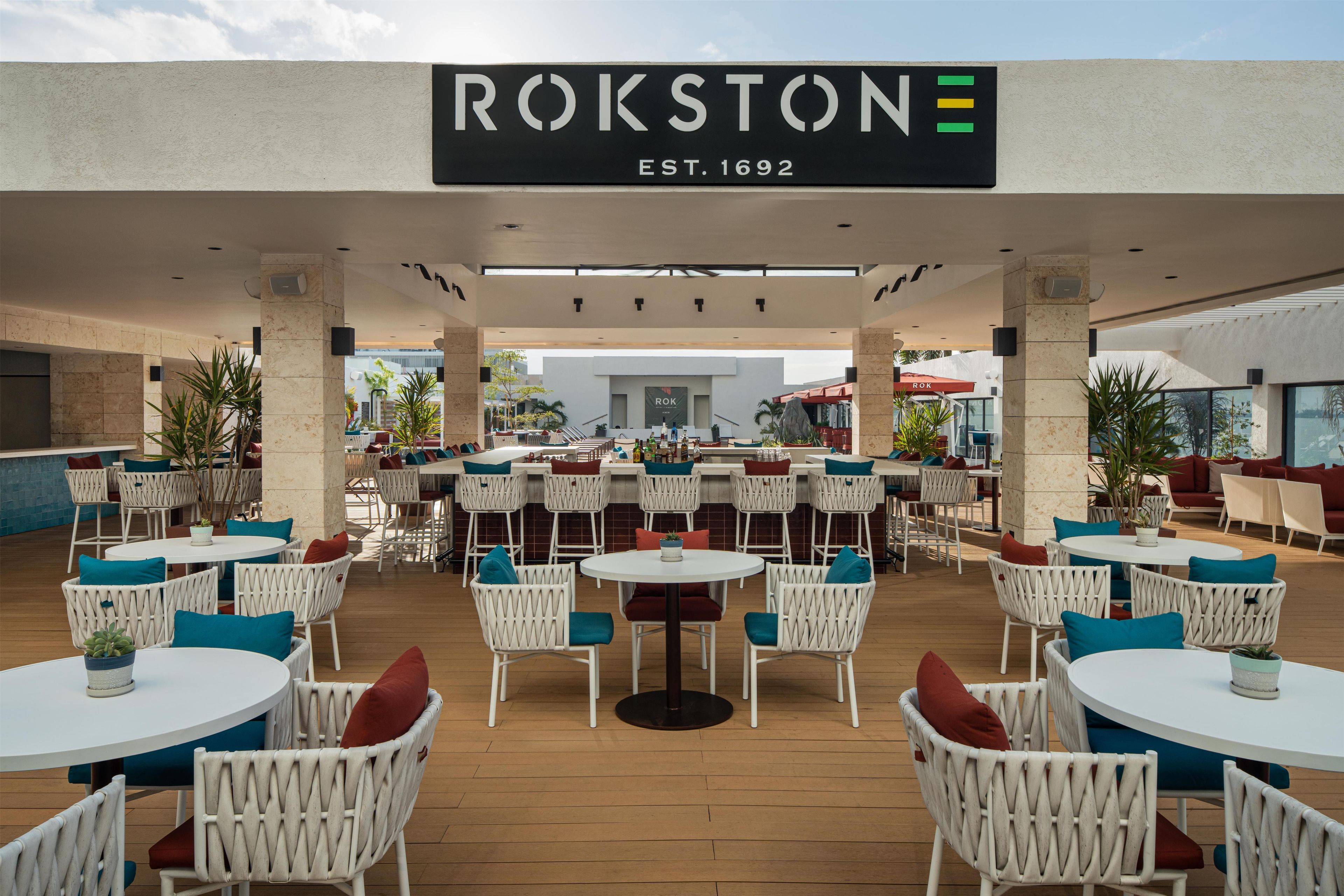 An outdoor seating area with white wicker chairs and tables, a sign reading 'ROKSTON EST. 1692', and a view of the building in the background.