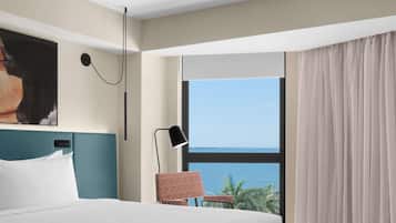 Habitación de hotel con una cama grande, una silla, una lámpara de escritorio, un cuadro en la pared y vista al mar a través de una ventana.