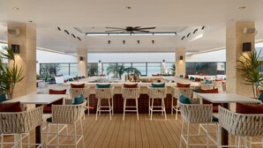 A spacious outdoor dining area with white wicker chairs and tables, a central bar, and large windows offering views of the ocean.