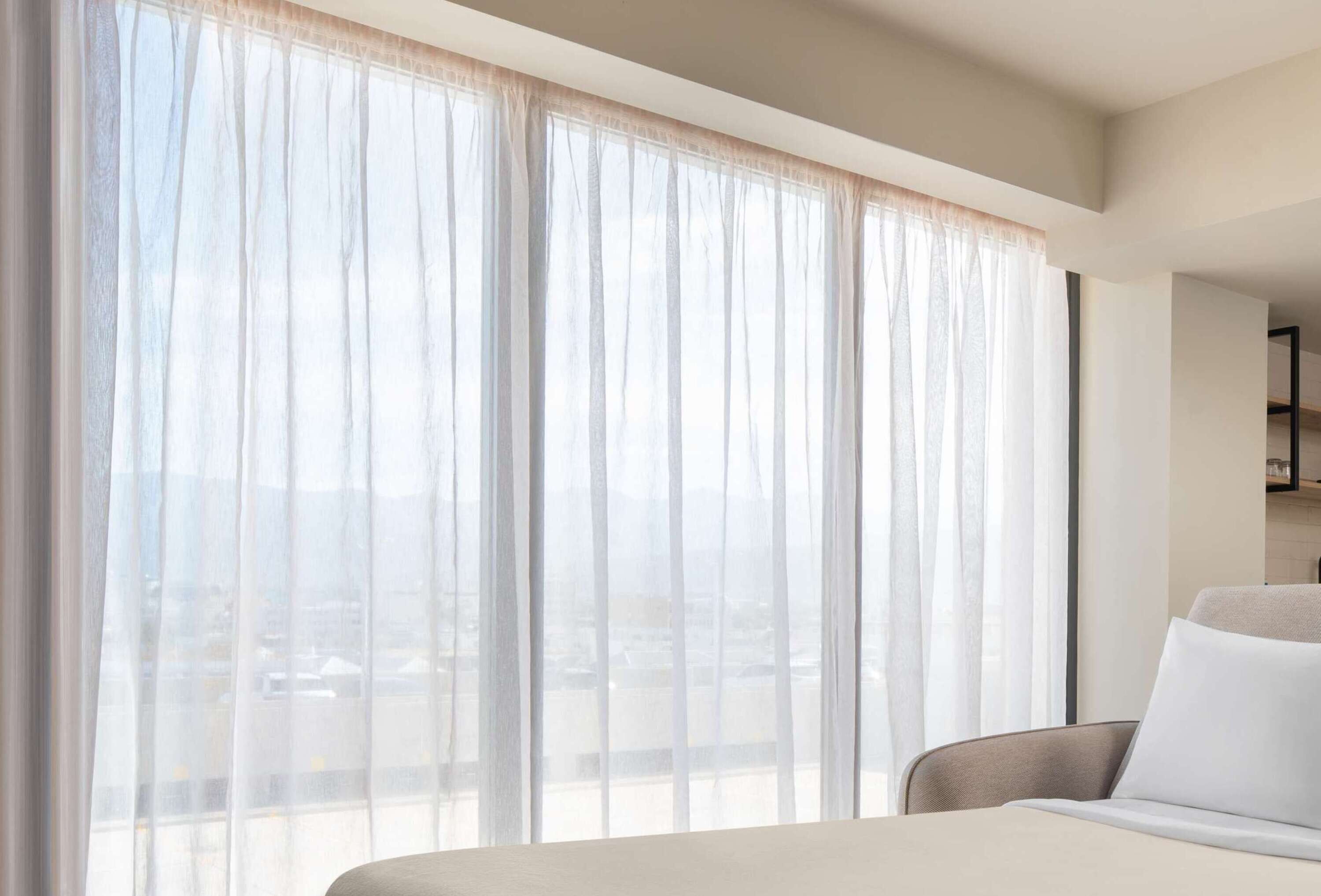 A hotel room with a bed, a chair, and large windows with sheer curtains.