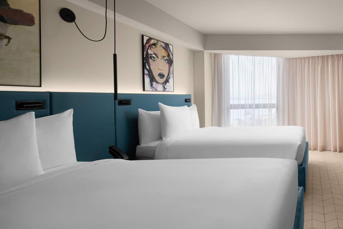 A hotel room with two beds, a large window with sheer curtains, and a painting on the wall.