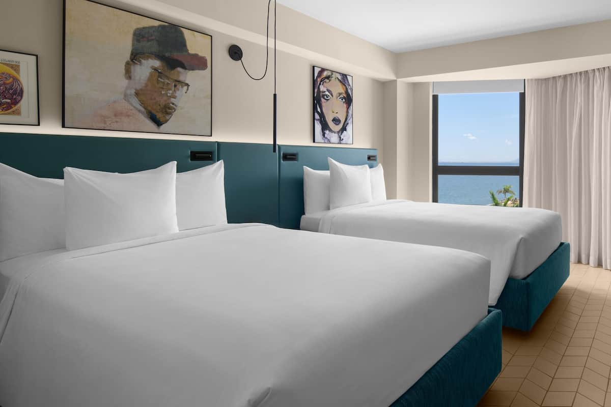 A hotel room with two beds, a large window with a view, and artwork on the walls.