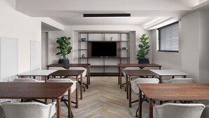 A conference room with a large flat-screen TV, wooden tables, white chairs, and potted plants.