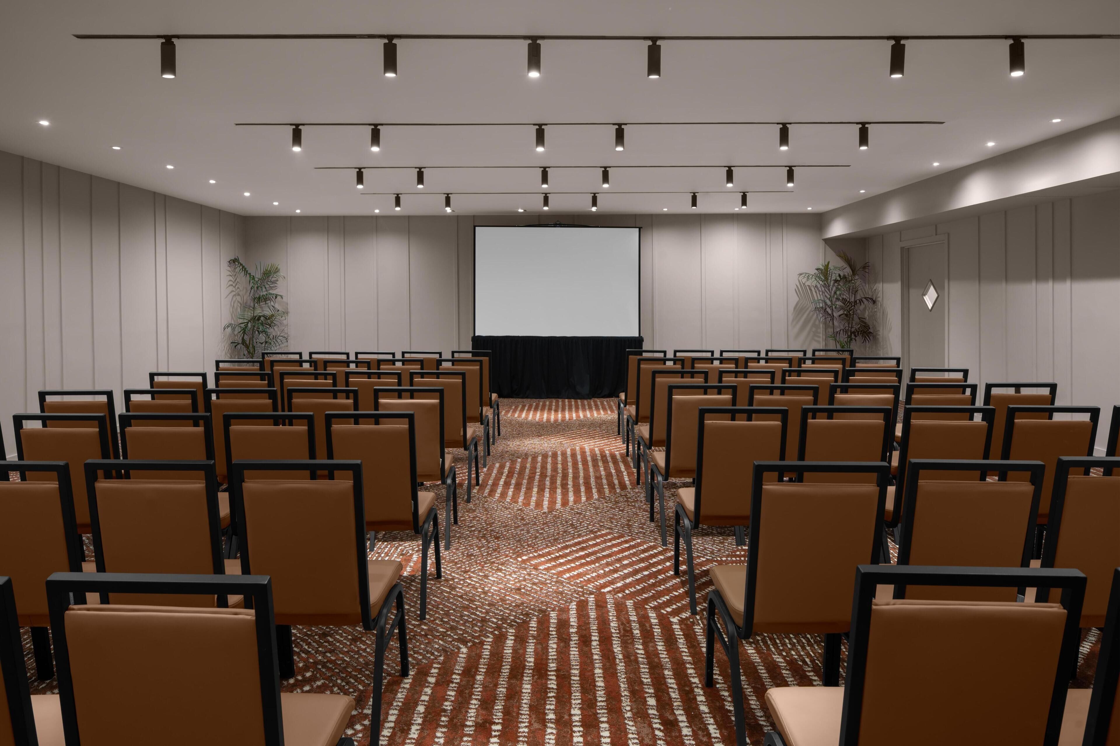 Un salón de conferencias con filas de sillas, una pantalla grande y luces en el techo.