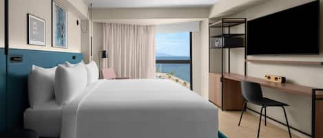 A hotel room with a bed, a desk, a chair, a TV, and a view of the sea.