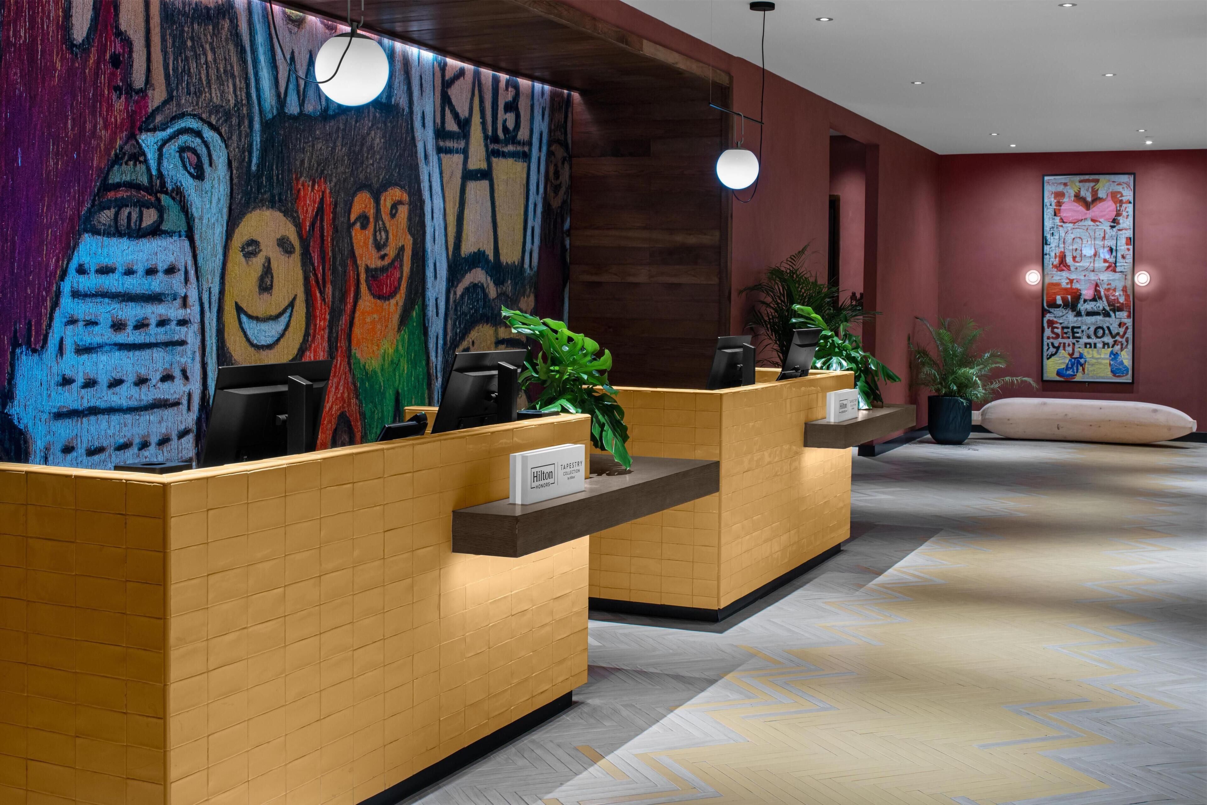 A hotel reception area with two reception desks, a colorful abstract mural, and a modern light fixture.