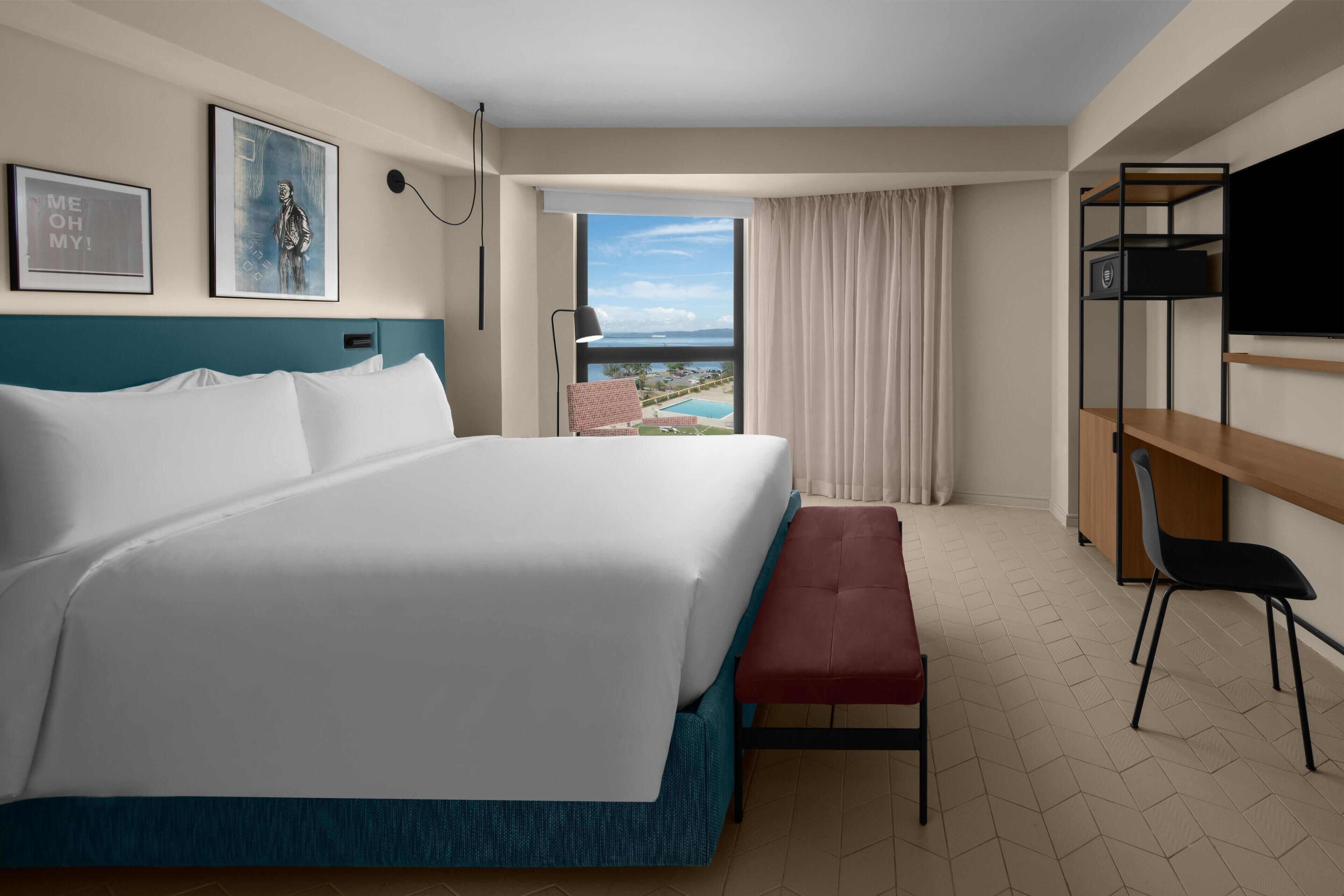 A hotel room with a large bed, a desk with a TV, a chair, and a view of a pool and landscape.