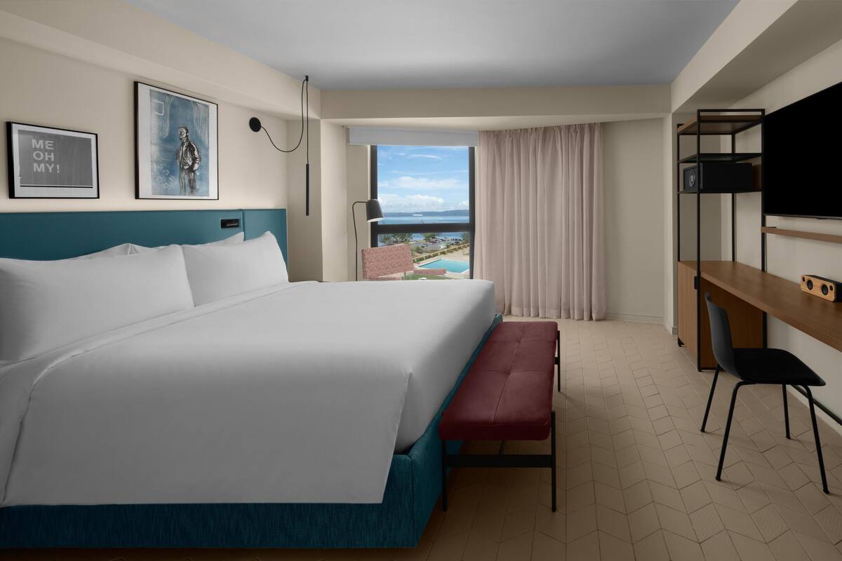 A hotel room with a large bed, a desk, a chair, a bench, a TV, and a view of a pool and landscape.