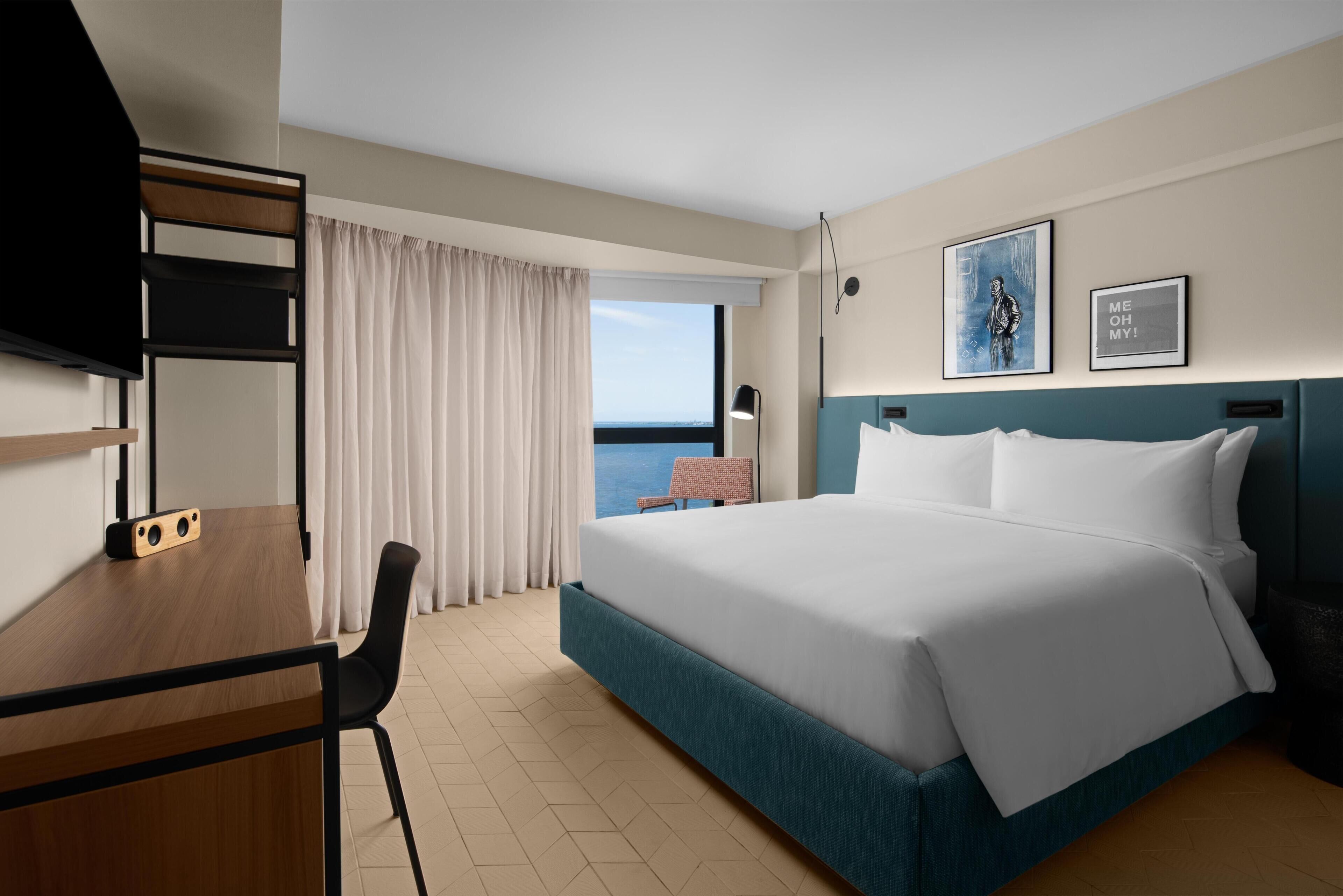 A hotel room with a bed, a desk, a chair, a television, and a view of the sea.