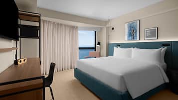 A hotel room with a bed, a desk, a chair, a television, and a view of the sea.